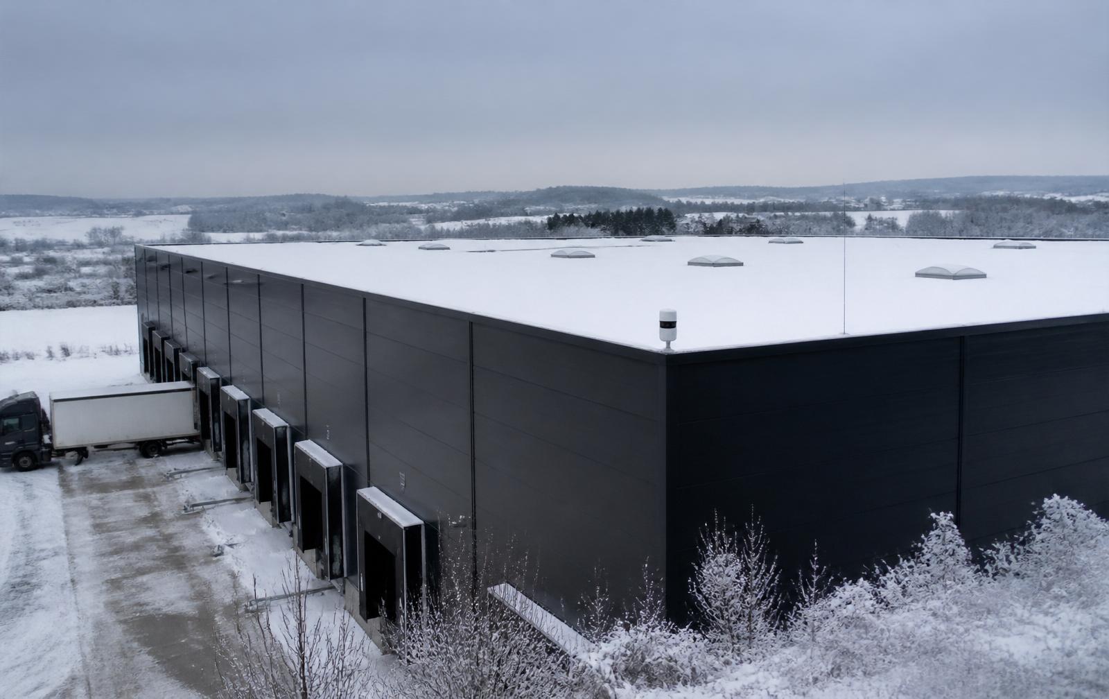 Außenansicht eines modernen Logistikzentrums mit schneebedecktem Flachdach, ausgestattet mit NevoSens Lasermessgeräten zur Durchbiegungsüberwachung